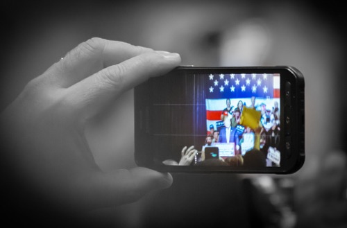 Trump on cell phone B&W A black and white hand holds up a cell phone to photograph President Trump