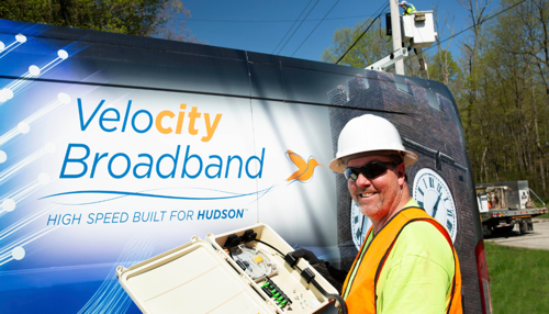 A city broadband employee stands next to Velocity Broadband van. He is smiling and holding a splice case