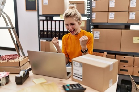 A happy business owner pumps her fist in excitement while looking at her laptop in front of a rack of cardboard boxes