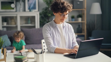 A mom works at home on computer with her child seated on couch in background