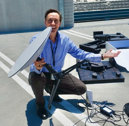 A man crouches with a smile on his face while holding a Starlink disk in his right hand amid installation equipment in a parking lot