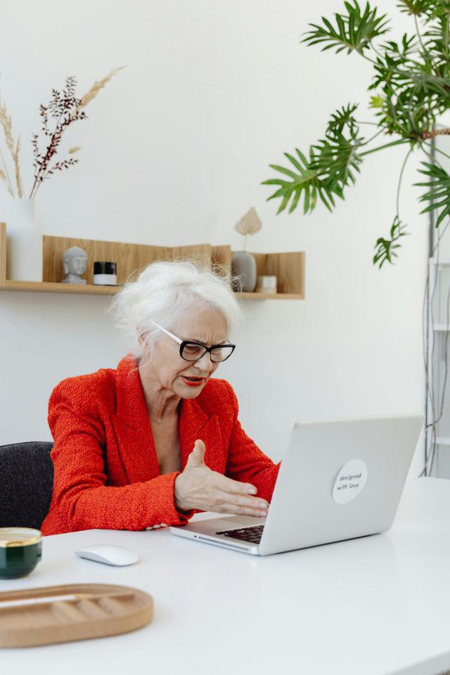An older lady wearing a red suit jacket looks distressingly at the laptop in front of her, hand motioning to the screen