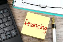 A yellow sticky on a desk next to computer keyboard reads: "financing"