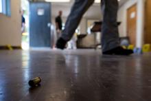 A bullet shell casing lays on linoleum floor in foreground with a pair of investigator legs walking by in the background