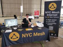 A volunteer sits behind a NYC Mesh table at a Maker Fair in NYC 