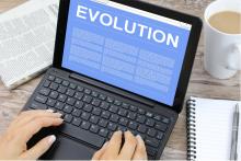A laptop on a desk is open next to a cup of coffee and a notebook with the word "Evolution" displayed on screen as a woman's hands rests on keyboard